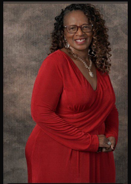 A smiling woman in a red dress with glasses and curly hair.